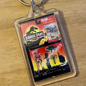 Może przedstawiać: Przezroczysty plastikowy brelok z kartą Jurassic Park przedstawiającą Ellie Sattler, małego dinozaura i zieloną roślinę. Karta zawiera tekst "Ellie Sattler -  Finding the Missing Herbivore!" i "Jurassic Park - Movie Collector Card".