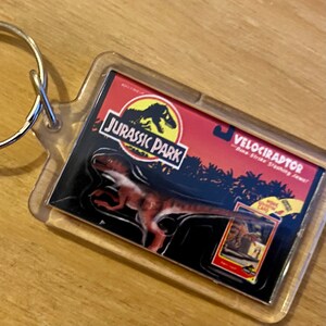 Może przedstawiać: Przezroczysty plastikowy brelok z czerwono-czarno-żółtym logo Jurassic Park i obrazem welociraptora. Tekst na breloku brzmi "Jurassic Park" i "Velociraptor - Dino Strike Striking Jaws!"