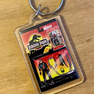 Może przedstawiać: Przezroczysty plastikowy brelok z figurką akcji Jurassic Park Dennisa Nedry z filmu. Figurka trzyma pistolet uspokajający i ma na sobie zieloną koszulę i czarne spodnie. Brelok zawiera również małą kartę z logo Jurassic Park i tekstem "Dennis Nedry - Tranq-Spray Gun - Dino-Damage Arm".