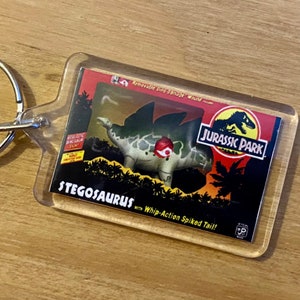 Może przedstawiać: Brelok z przezroczystego plastiku z zabawką Stegosaurus z Jurassic Park. Zabawka jest zielona i szara z czerwonym siodłem i ogonem z kolcami z działaniem bicza. Brelok zawiera tekst "Removable Dino-Damage Wound", "Jurassic Park", "Stegosaurus" i "Whip-Action Spiked Tail!"