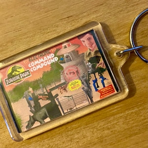 Może przedstawiać: Przezroczysty plastikowy brelok z reklamą zabawki Jurassic Park Electronic Command Compound. Na zdjęciu widać chłopca bawiącego się zabawką, która zawiera wieżę, bramę i dinozaura.