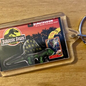 Może przedstawiać: Przezroczysty plastikowy brelok z logo Jurassic Park i obrazkiem dinozaura Dimetrodon. Brelok zawiera napis "Dino-Strike Clamping Jaws!" i małą kartkę z obrazkiem dinozaura.