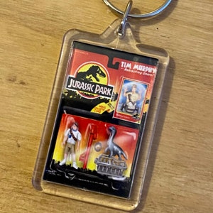 Może przedstawiać: Przezroczysty plastikowy brelok z motywem Jurassic Park. Brelok zawiera małą plastikową figurkę dinozaura, małą plastikową figurkę postaci z filmu oraz kartę z tekstem "Jurassic Park" i "Tim Murphy - Retracting Snare!"