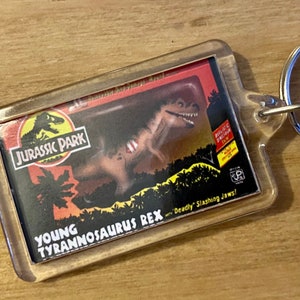 Może przedstawiać: Przezroczysty plastikowy brelok do kluczy z czerwoną i żółtą kartą Jurassic Park przedstawiającą młodego Tyrannosaurus Rexa z tekstem "Young Tyrannosaurus Rex with Deadly Slashing Jaws!"