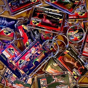 Może przedstawiać: Zbiór kolorowych breloków z różnymi postaciami i logo z Jurassic Park. Breloki wykonane są z przezroczystego plastiku i przedstawiają obrazy dinozaurów, pojazdów i tekstu z filmu.