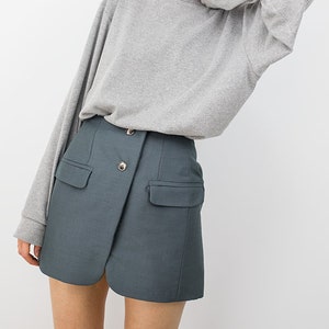 May include: A woman wearing a gray long-sleeved sweater and a dark teal skirt with a button closure and pockets.