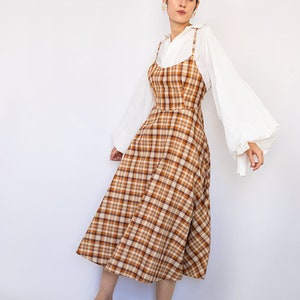 Pode incluir: Uma mulher vestindo um vestido midi xadrez marrom e branco com uma camisa branca por baixo. A camisa tem mangas compridas e bufantes. Ela est&aacute; usando sand&aacute;lias verdes.
