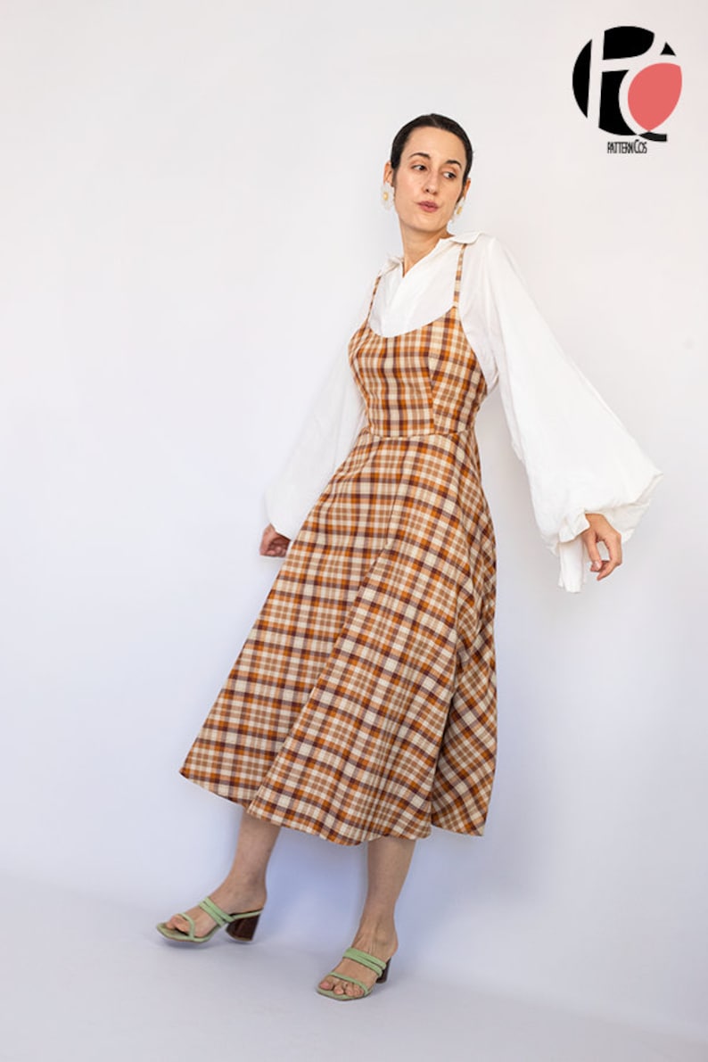 Pode incluir: Uma mulher vestindo um vestido midi xadrez marrom e branco com uma camisa branca por baixo. A camisa tem mangas compridas e bufantes. Ela est&aacute; usando sand&aacute;lias verdes.