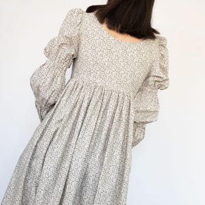 May include: A long, light-colored dress with a floral pattern. The dress features long sleeves with puffed shoulders and a gathered skirt. The model is seen from the back, with the dress's details clearly visible against a white background.