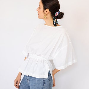 May include: A white, loose-fitting blouse with a gathered waist and a side slit. The blouse has short, wide sleeves and a round neckline. The model is wearing blue jeans.