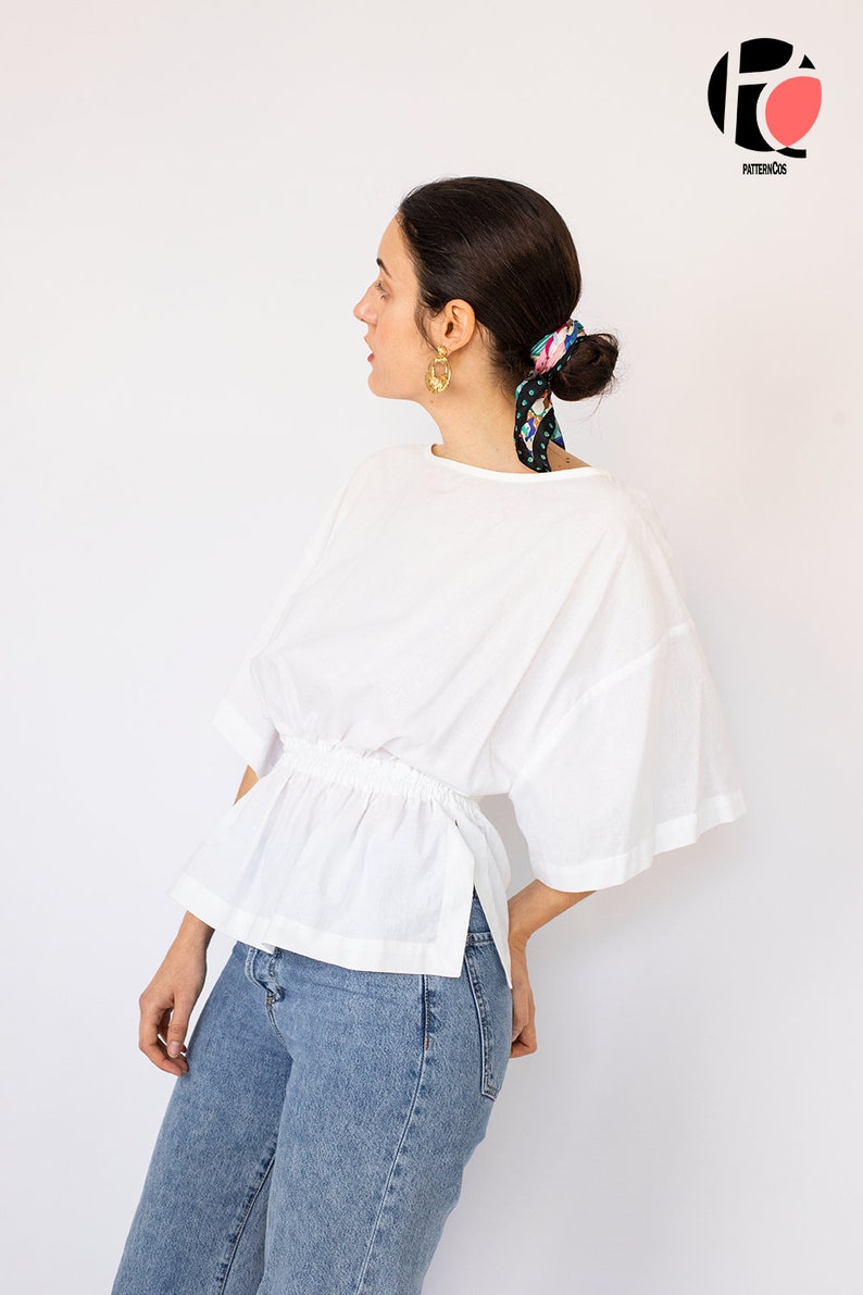 May include: A white, loose-fitting blouse with a gathered waist and a side slit. The blouse has short, wide sleeves and a round neckline. The model is wearing blue jeans.