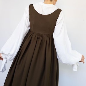 May include: A woman wearing a dark brown dress with a white blouse underneath. The dress has a scoop neckline and a gathered skirt. The white blouse has long, billowy sleeves. The logo for PatternCos is in the upper right corner.