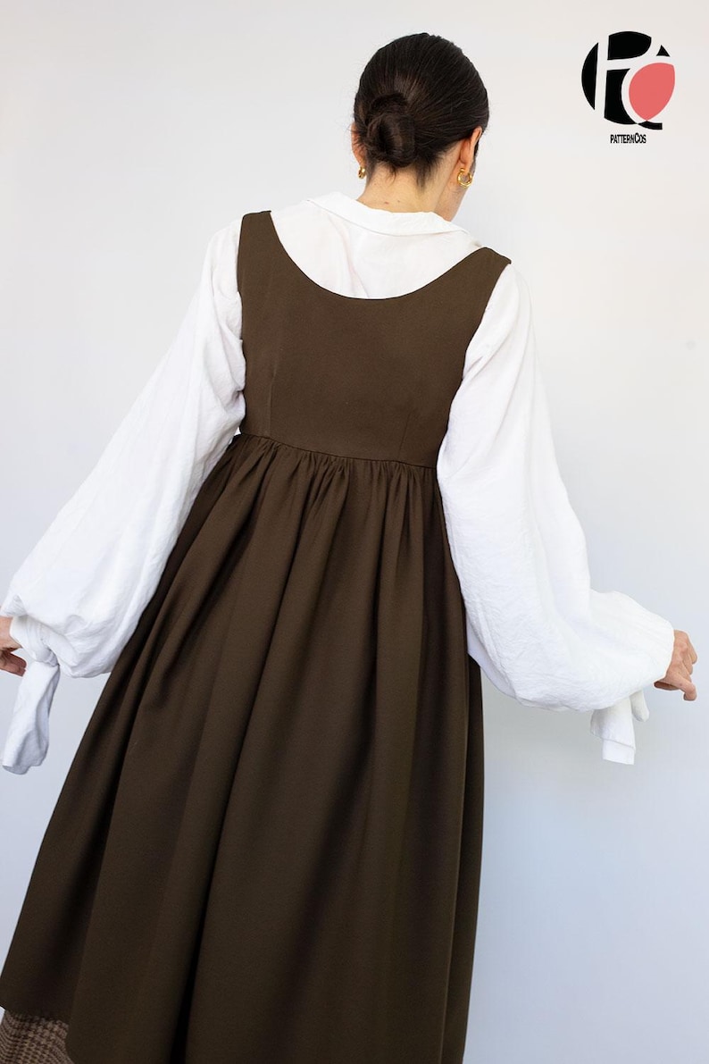 May include: A woman wearing a dark brown dress with a white blouse underneath. The dress has a scoop neckline and a gathered skirt. The white blouse has long, billowy sleeves. The logo for PatternCos is in the upper right corner.