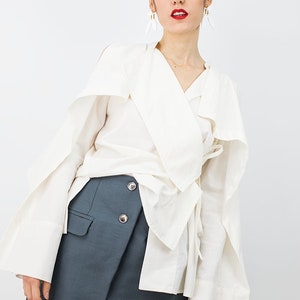 May include: A white long-sleeved blouse with a wrap-style neckline and a tie at the waist. The blouse has wide, flowing sleeves. The model is also wearing a dark gray skirt with a wrap-style front and three buttons.