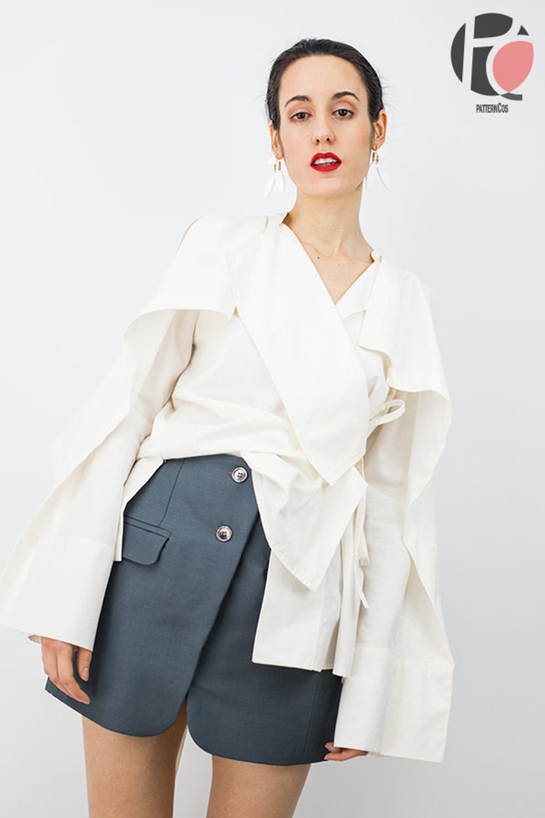 May include: A white long-sleeved blouse with a wrap-style neckline and a tie at the waist. The blouse has wide, flowing sleeves. The model is also wearing a dark gray skirt with a wrap-style front and three buttons.