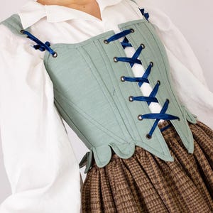 May include: A light green corset with dark blue ribbon lacing. The corset is worn over a white blouse and a brown plaid skirt. The corset has a scalloped edge and is part of a historical costume.