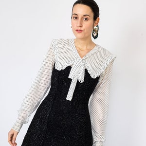 K&ouml;nnte beinhalten: Ein schwarzes Tweed-Minikleid mit einer wei&szlig;en Bluse mit Polka Dots, die einen gro&szlig;en Kragen und eine Schleife aufweist. Das Kleid hat lange &Auml;rmel und ein figurbetontes Oberteil. Das Model tr&auml;gt gro&szlig;e Ohrringe und hat dunkles Haar.