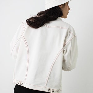 Pu&ograve; includere: Una giacca di jeans bianca con cuciture rosse. La giacca ha una chiusura con bottoni e una vestibilit&agrave; rilassata. La modella indossa la giacca con pantaloncini da ciclista neri e un cappello a secchiello bianco.