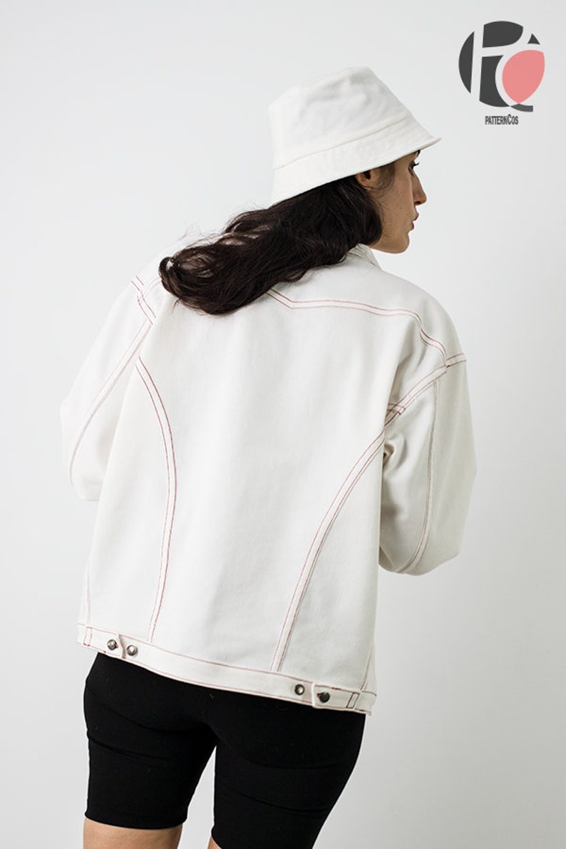 Pu&ograve; includere: Una giacca di jeans bianca con cuciture rosse. La giacca ha una chiusura con bottoni e una vestibilit&agrave; rilassata. La modella indossa la giacca con pantaloncini da ciclista neri e un cappello a secchiello bianco.