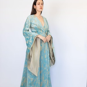 May include: A long, flowing teal dress with gold accents and wide sleeves. The dress features a wrap-style front and a matching sash. The fabric has a soft, velvety texture. The dress is suitable for special occasions.