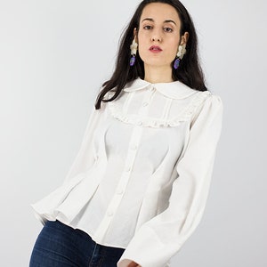 May include: A white button-up blouse with a ruffled collar and long sleeves. The blouse has a fitted bodice and a flared hem. The model is wearing blue jeans.