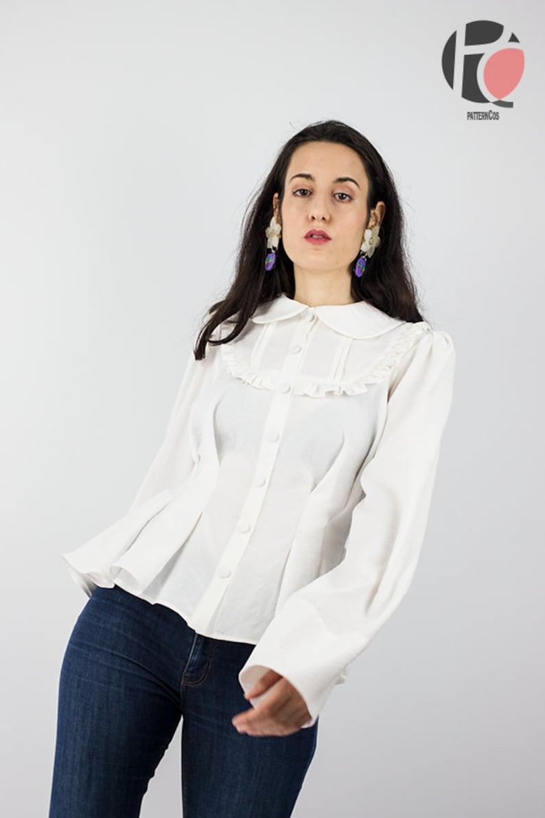 May include: A white button-up blouse with a ruffled collar and long sleeves. The blouse has a fitted bodice and a flared hem. The model is wearing blue jeans.