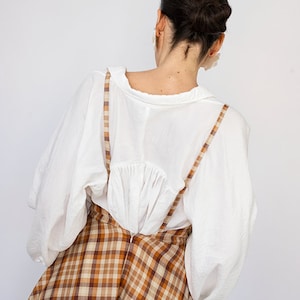 Pode incluir: Uma blusa branca de mangas compridas e uma saia xadrez marrom e bege com suspens&oacute;rios. A saia &eacute; de corte A e chega aos joelhos.