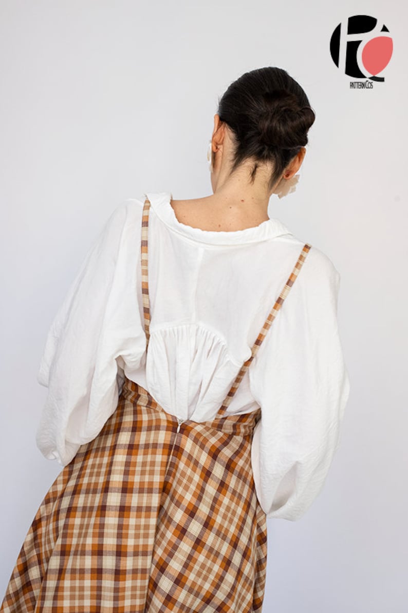 Pode incluir: Uma blusa branca de mangas compridas e uma saia xadrez marrom e bege com suspens&oacute;rios. A saia &eacute; de corte A e chega aos joelhos.