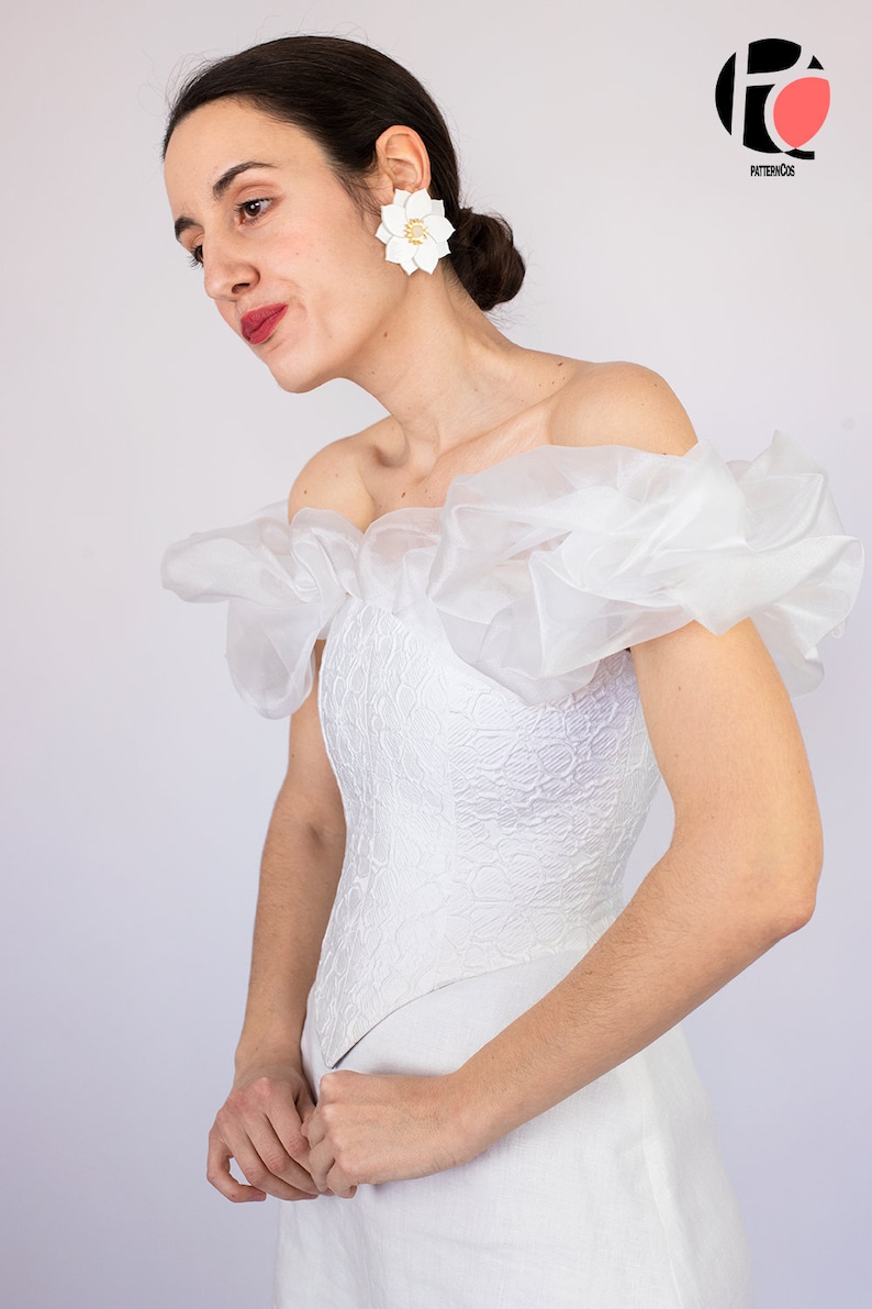 May include: A white off-the-shoulder dress with a lace bodice and a ruffled overlay. The dress has a fitted bodice and a flowing skirt. The model is wearing white flower earrings.
