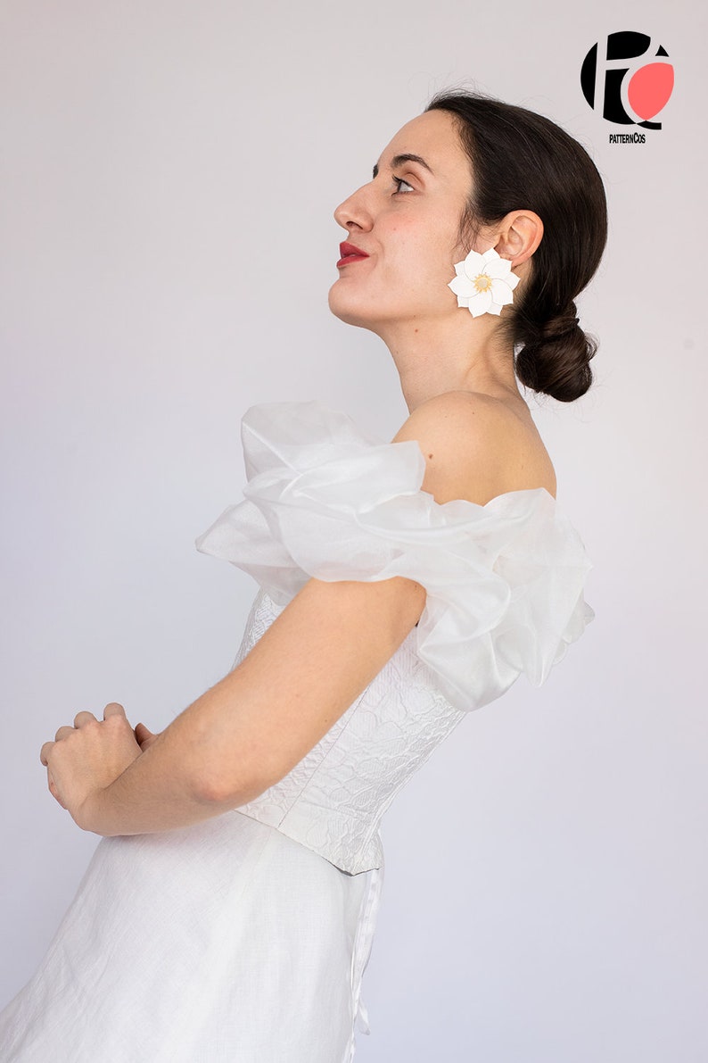 May include: A white off-the-shoulder dress with a lace bodice and a tulle overlay. The dress has a fitted bodice and a flowing skirt. The model is wearing large white flower earrings.