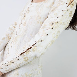 Peut inclure: Robe blanche en dentelle avec des broderies dor&eacute;es et des d&eacute;tails de la&ccedil;age sur les manches.