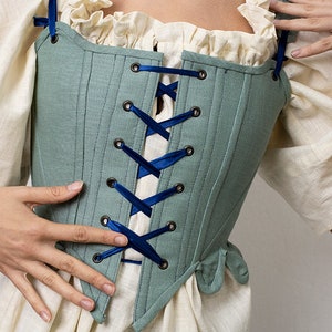 May include: A teal corset with dark blue ribbon lacing and eyelets. The corset is worn over a cream-colored blouse with ruffled trim. The garment has a structured, tailored appearance, suggesting historical or costume design.