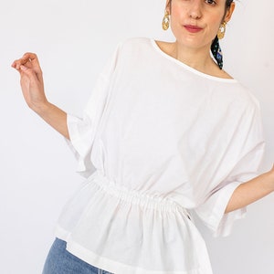 May include: A white, loose-fitting blouse with a gathered elastic waistband and 3/4 sleeves. The blouse is made of a lightweight fabric and has a relaxed fit.