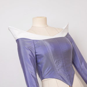 Peut inclure: Un corsage violet avec un col blanc et des manches longues. Le corsage est ajust&eacute; et a une taille pointue. Le tissu est brillant et a un l&eacute;ger &eacute;clat. Le corsage est sur un mannequin.