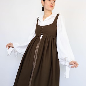 May include: A brown pinafore dress with a square neckline and button details, layered over a white blouse with voluminous sleeves. The dress is paired with a patterned skirt. The logo "PATTERNCOS" is in the top right corner.
