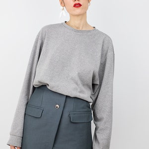 May include: A woman wearing a gray long-sleeved sweatshirt and a dark blue mini skirt with a single button closure and two pockets.