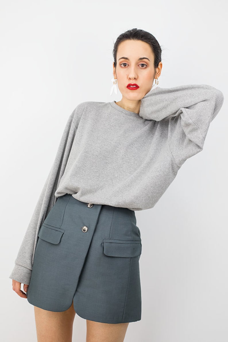 May include: A woman wearing a gray long-sleeved sweater and a dark teal skirt with a button closure and pockets.