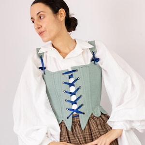 May include: A light green corset with blue ribbon lacing is worn over a white blouse with puffy sleeves. The corset has a square neckline and is paired with a brown plaid skirt. The image is set against a white background.