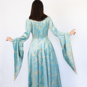 May include: A long, flowing dress in a light blue hue with a gold pattern. The dress features long sleeves with wide, flared cuffs. The model is facing away from the camera, showcasing the dress's design and silhouette. The dress is made of a soft, flowing fabric.