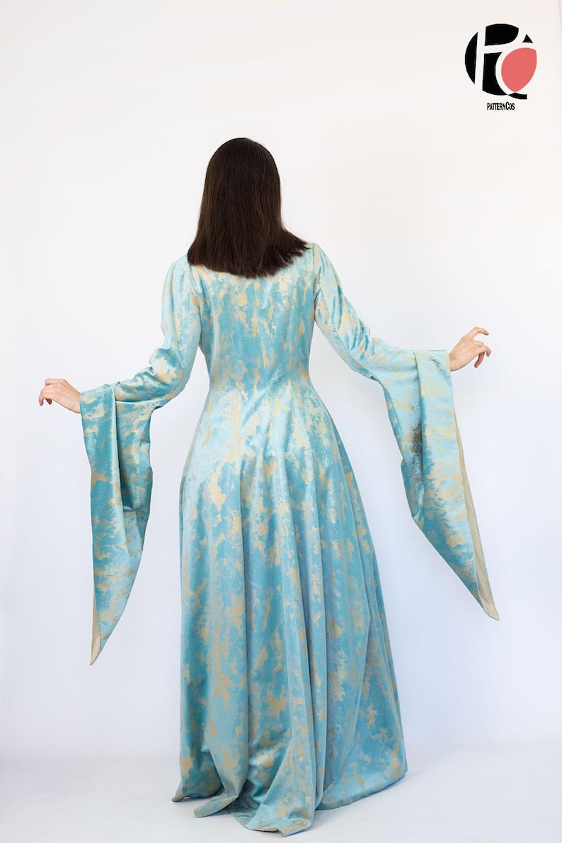 May include: A long, flowing dress in a light blue hue with a gold pattern. The dress features long sleeves with wide, flared cuffs. The model is facing away from the camera, showcasing the dress's design and silhouette. The dress is made of a soft, flowing fabric.