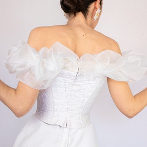 May include: White off-the-shoulder corset top with a lace-up back and ruffled sleeves. The top is made of a sheer fabric and has a fitted silhouette.