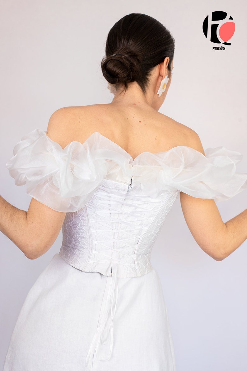 May include: White off-the-shoulder corset top with a lace-up back and ruffled sleeves. The top is made of a sheer fabric and has a fitted silhouette.