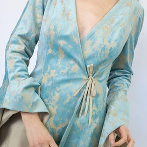 May include: A light blue wrap dress with long sleeves and a V-neck. The dress features a gold pattern and a tie closure at the waist. The sleeves have a flared design. The dress is made of a soft, flowing fabric.