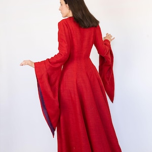 May include: A long, red dress with long sleeves and a flared skirt. The sleeves have a wide, bell-like shape with a purple lining. The dress is made of a textured fabric and has a high neckline. The dress is a solid red color.
