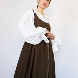 May include: A woman wearing a layered outfit. A white blouse with voluminous sleeves is paired with a dark brown dress. The dress features a button-down front and a long, flowing skirt. The outfit has a vintage or historical aesthetic.