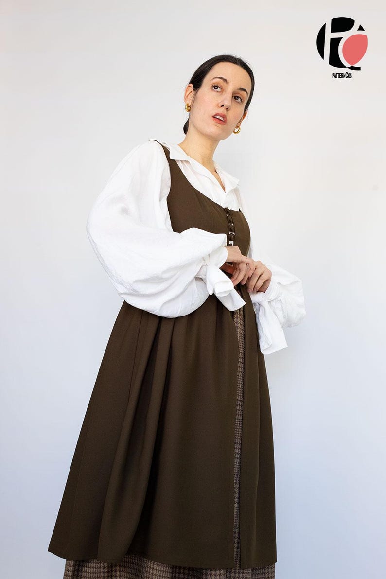 May include: A woman wearing a layered outfit. A white blouse with voluminous sleeves is paired with a dark brown dress. The dress features a button-down front and a long, flowing skirt. The outfit has a vintage or historical aesthetic.