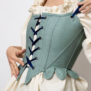 May include: A teal corset with dark blue ribbon lacing. The corset has a scalloped hem and is worn over a cream-colored blouse with a ruffled neckline. The logo "PatternCos" is visible in the upper left corner.