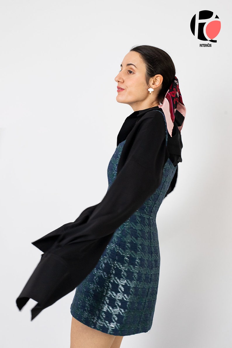K&ouml;nnte beinhalten: Eine Frau tr&auml;gt ein schwarzes Langarm-Oberteil &uuml;ber einem blau-gr&uuml;nen Mini-Kleid mit Hahnentrittmuster. Ein gemustertes Halstuch und wei&szlig;e Ohrringe vervollst&auml;ndigen den Look. Das Bild ist vor einem wei&szlig;en Hintergrund aufgenommen.