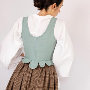 May include: A light green corset with a scalloped hem is worn over a white blouse with puffy sleeves. A brown plaid skirt completes the historical outfit. The image features the PatternCos logo in the upper right corner.