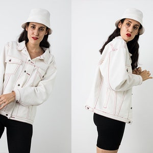 Pu&ograve; includere: Una giacca di jeans bianca con cuciture rosse. La giacca ha una chiusura con bottoni e due tasche sul davanti. La modella indossa pantaloncini neri e un cappello a secchiello bianco.